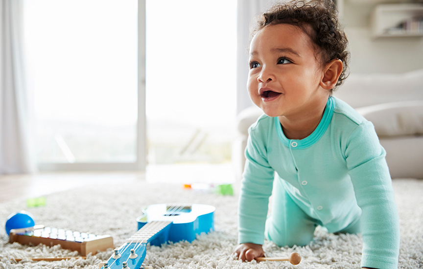 A baby in a mint green onesie smiles and crawls on a carpet scattered with toys.