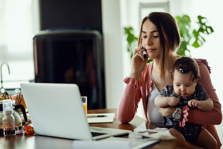 Woman sitting at table holding baby with laptop open 